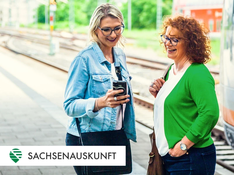 Zwei Frauen stehen am Bahnsteig und blicken in ein Mobiltelefon