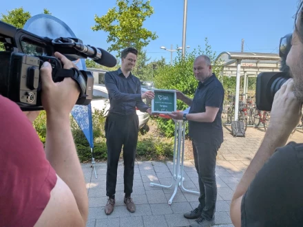 Kameras filmen ene Preisübergabe, zwei Männer halten eine Preisplakette hoch