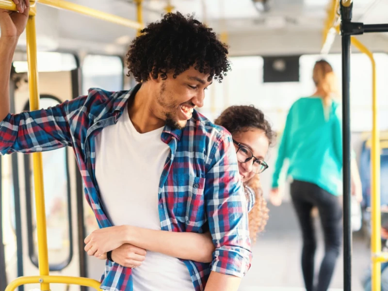eine junge Frau umarmt von hinten einen jungen Mann in einer Tram, beide lächeln